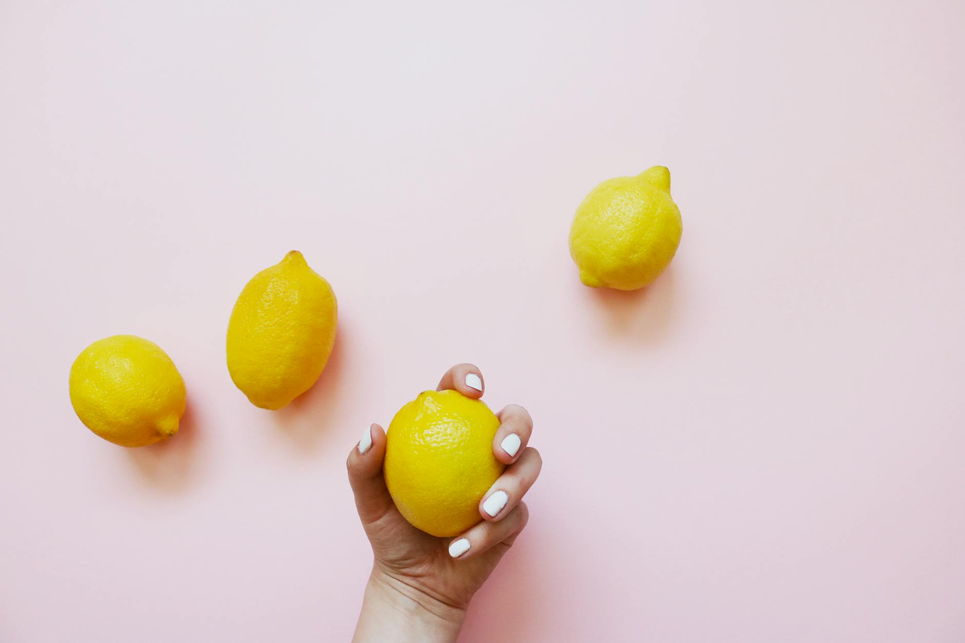 Una mano con smalto bianco che tiene un limone su uno sfondo rosa tenero, circondata da tre limoni aggiuntivi.