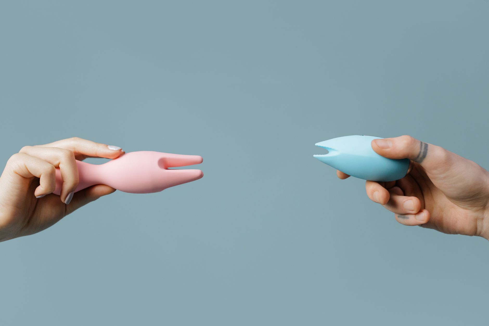 Due mani che tengono vibratori in silicone rosa e blu su uno sfondo pastello.