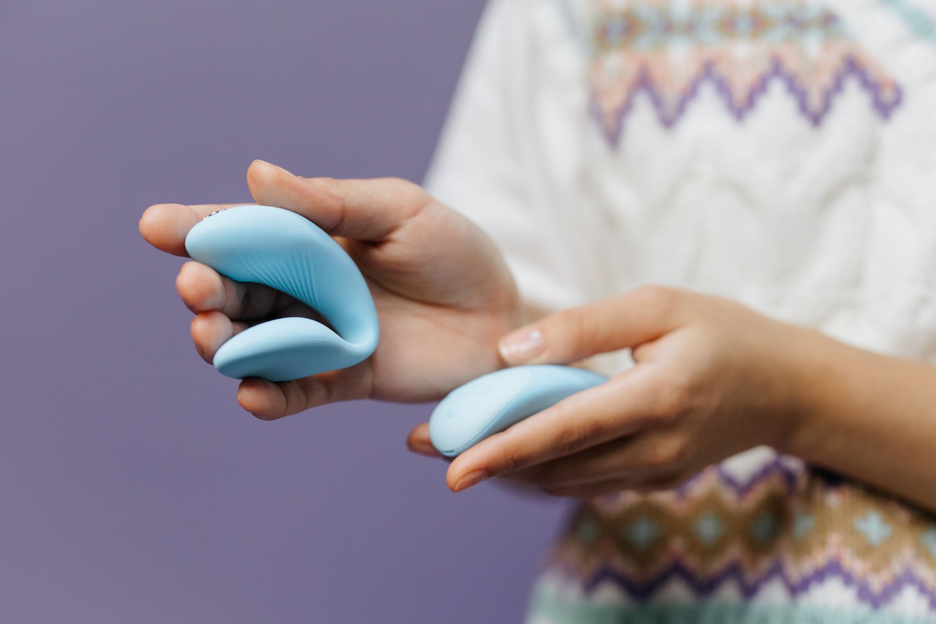 Two hands holding pink and blue silicone vibrators against a pastel background.